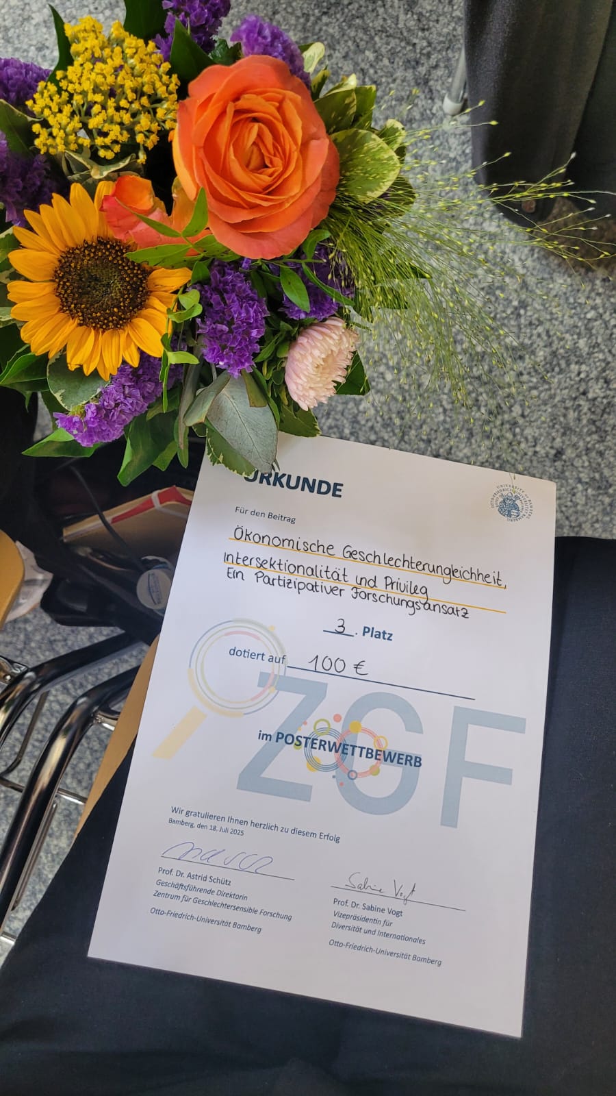 Am linken Bildrand ein Blumenstrauß und im Bild eine Urkunde für den dritten Presi des Posterwettbewerbes des Zentrum für Geschlchetersensible Forschung der Uni Bamberg